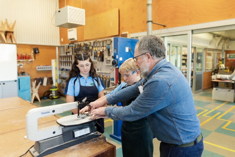 Male teacher with two students in woodwork workshop standing at a bench saw, teacher showing students how to hold wood when using saw