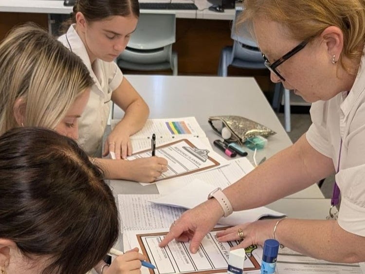 Female teacher wearing glasses with three students working on writing tasks, teacher at students desk pointing to task and interacting with students