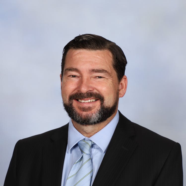 portrait photo of Principal Paul Baxter with dark hair and a beard and moustache, smiling at camera wearing a blazer and striped tie